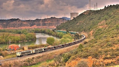 A train traveling through a hilly landscape with a river, power lines, and a large water tower.