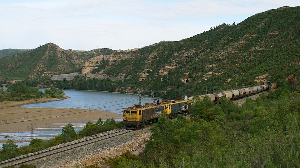 A train on tracks with a river and hills in the background.