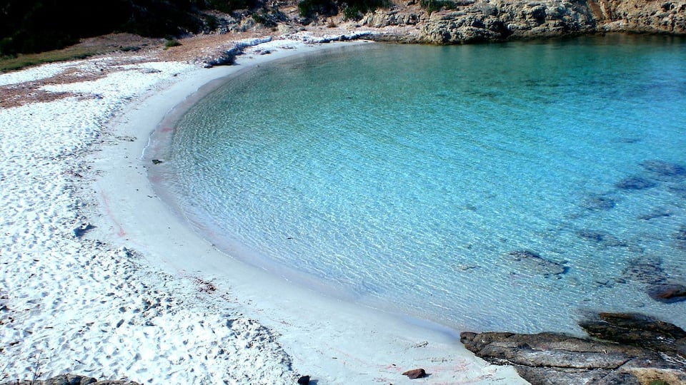 A clear, curved sandy beach with rocky outcrops and sparse vegetation.