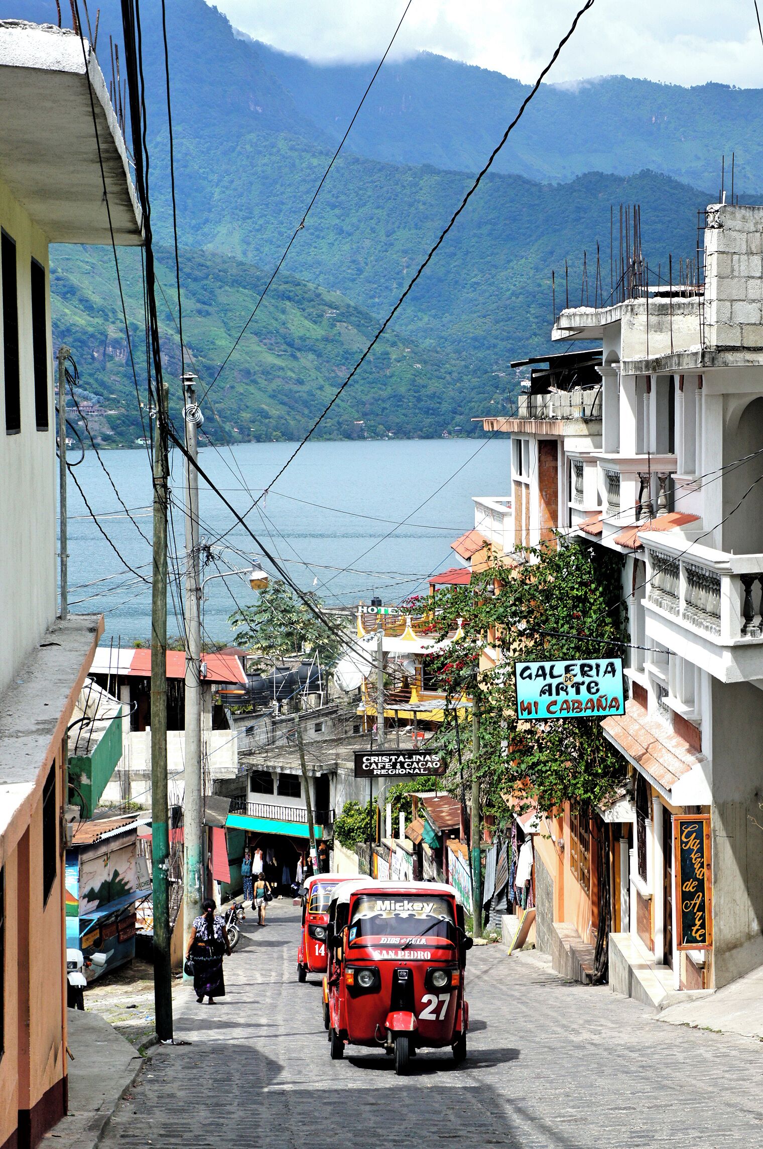 A street with a view of a lake and mountains in the background. There are buildings, a sign for 'Galeria de Arte de Mi Cabaña', and a red vehicle with 'Mickey' on it.