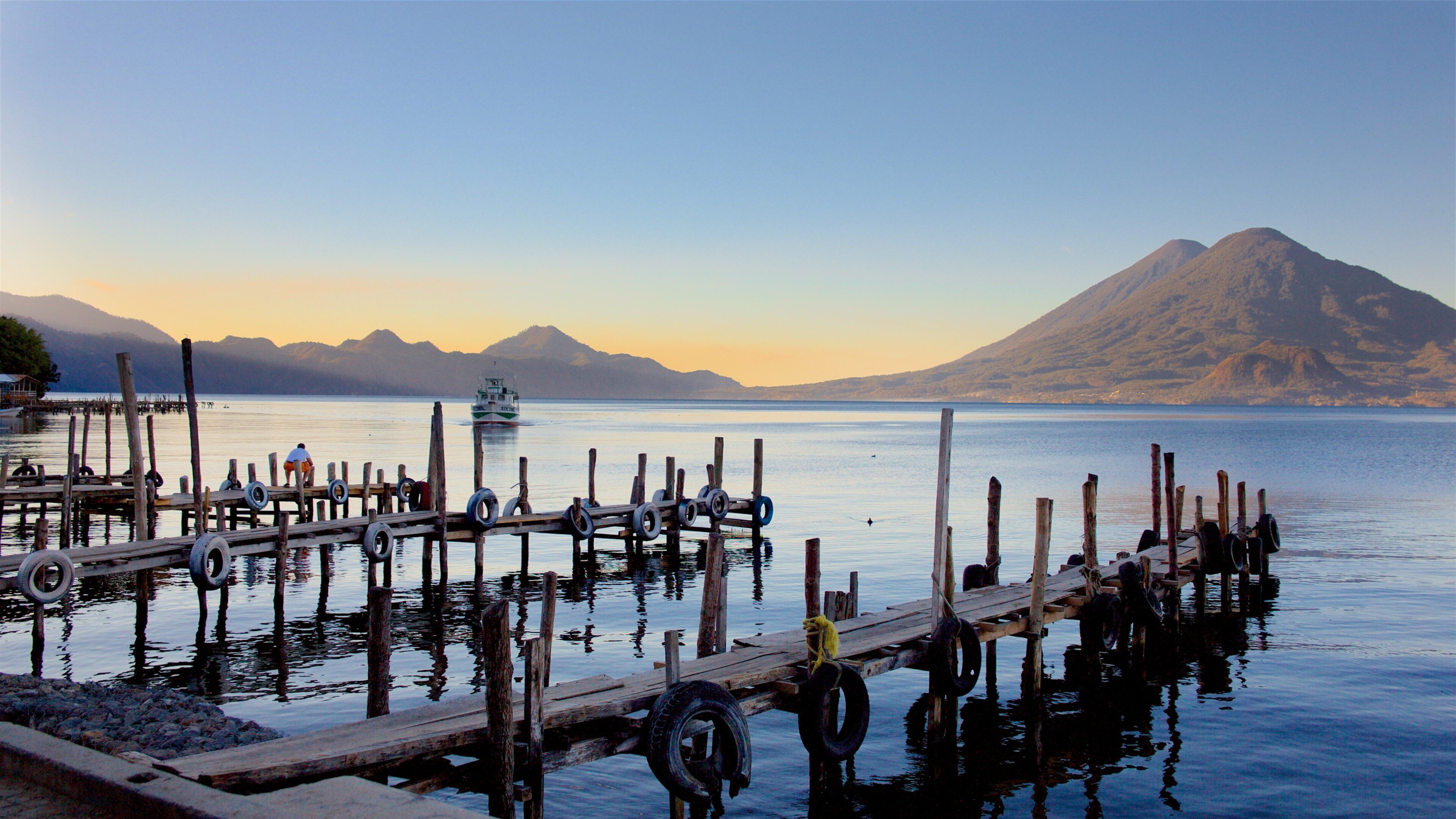 A wooden pier with tires and a boat in the water, a mountain in the background, and a clear sky.