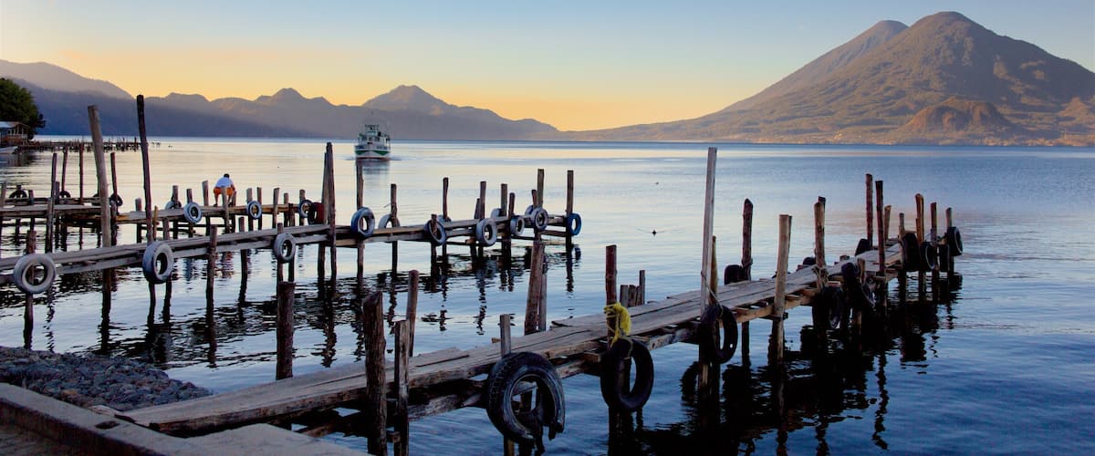 A wooden pier with tires and a boat in the water, a mountain in the background, and a clear sky.
