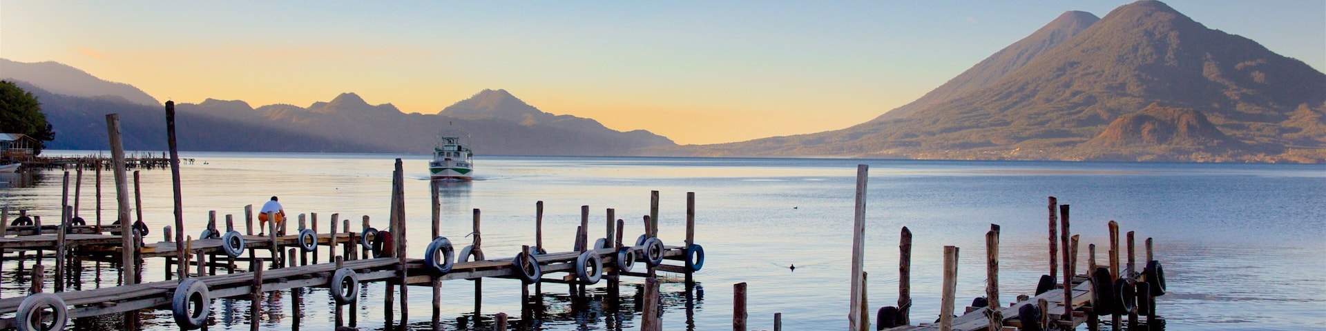 A wooden pier with tires and a boat in the water, a mountain in the background, and a clear sky.