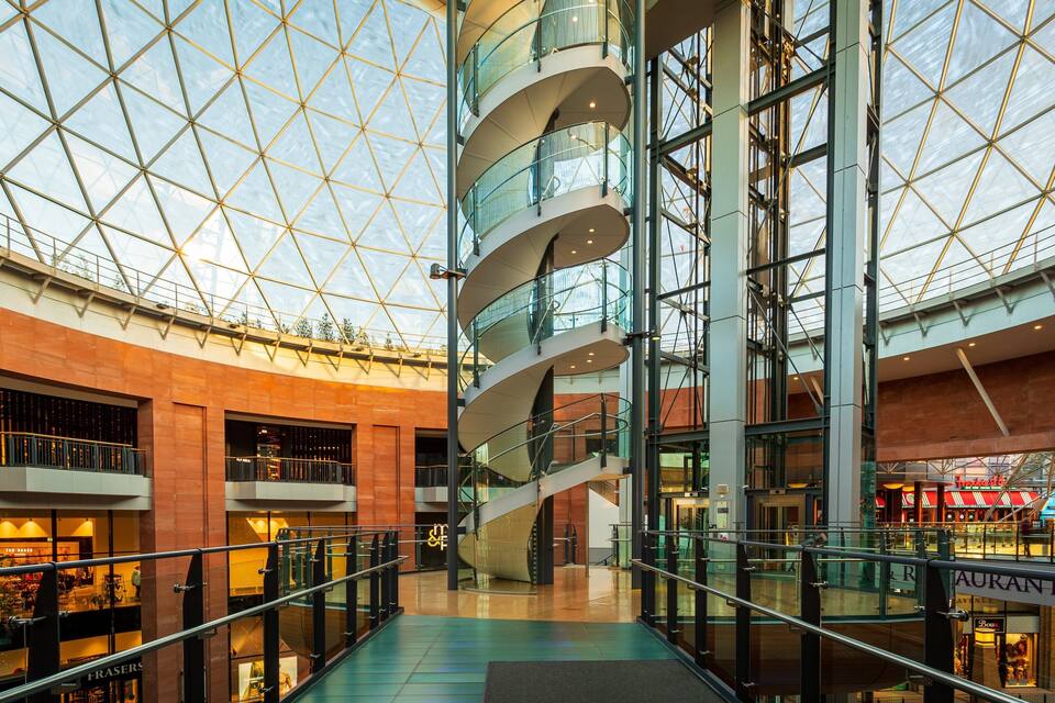 Um moderno centro comercial com cúpula de vidro, vários andares e uma escadaria espiral central.