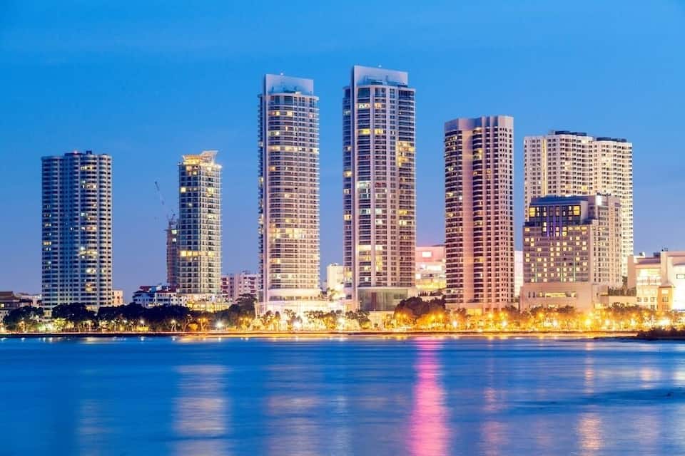 A city skyline with tall buildings along a waterfront at dusk.