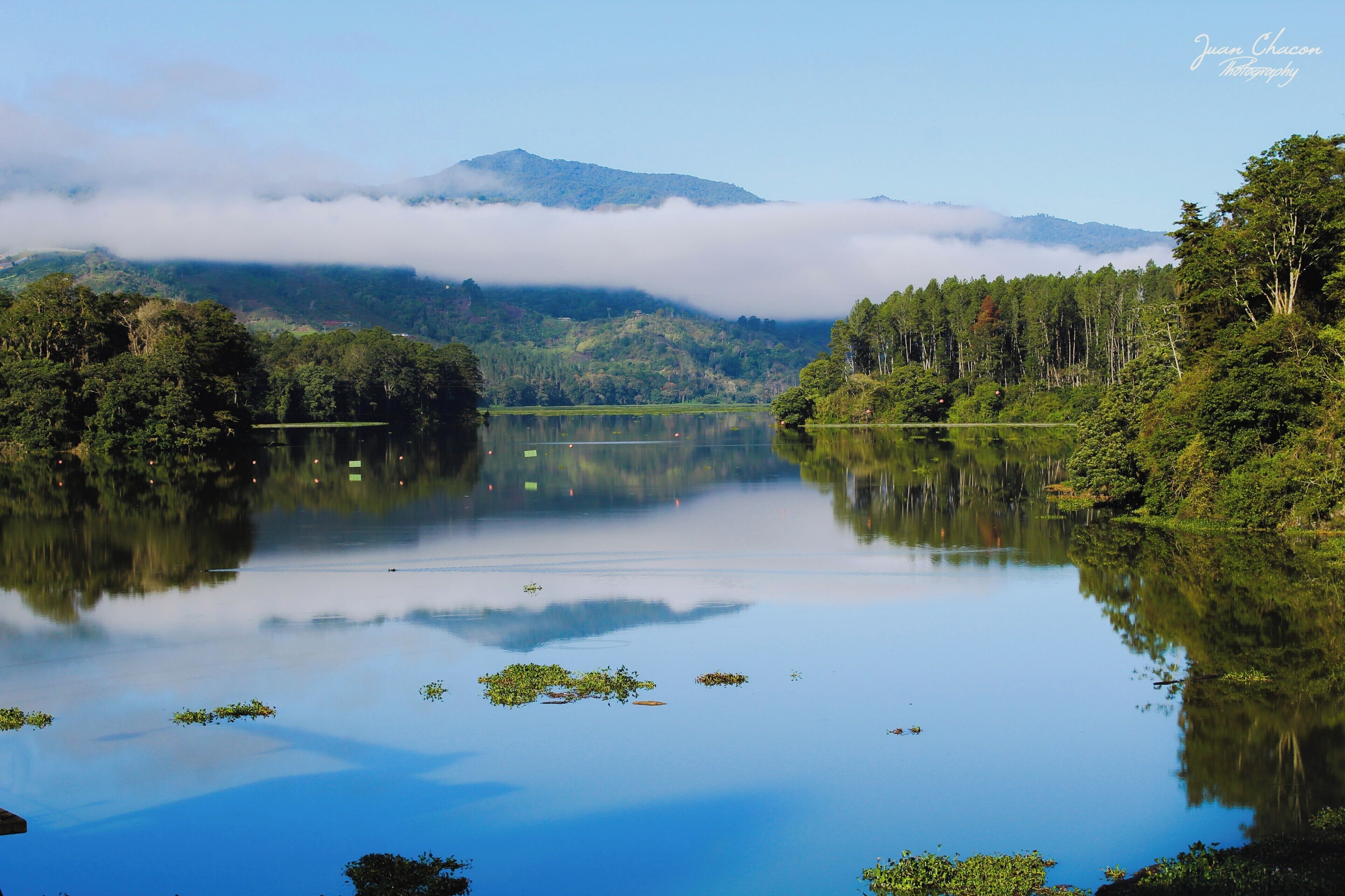 A calm lake surrounded by dense forest and mountains, with a mist-covered peak in the background.