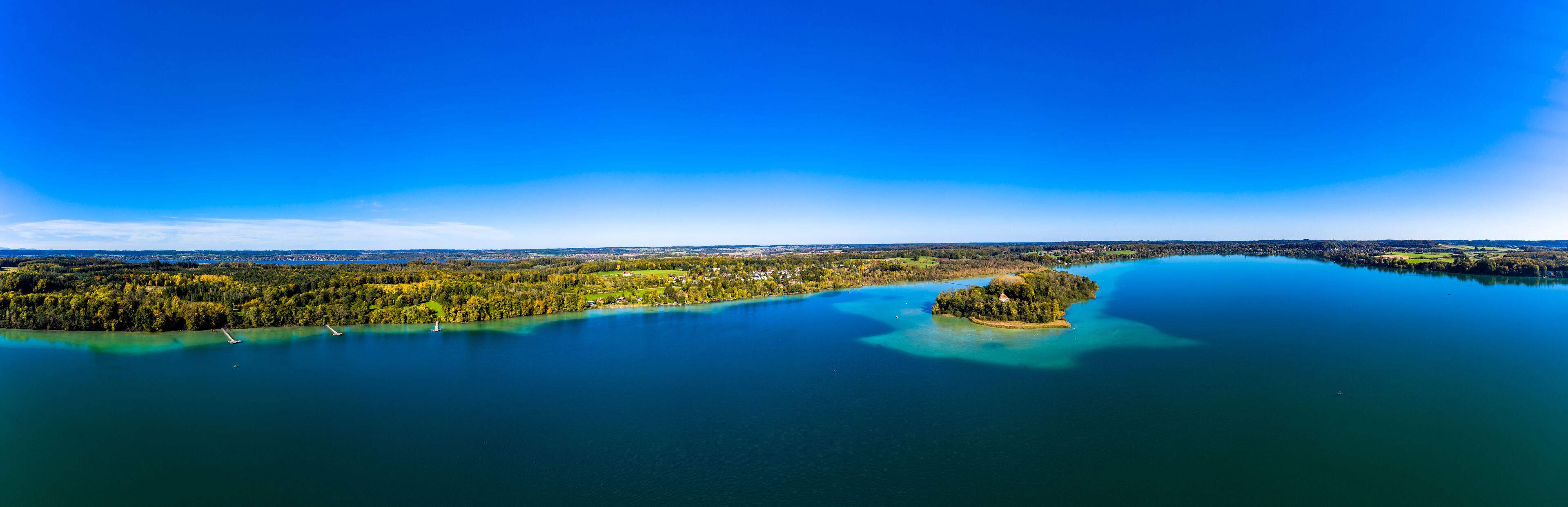 A panoramic view of a lake surrounded by dense forests and a small island with a lighthouse.