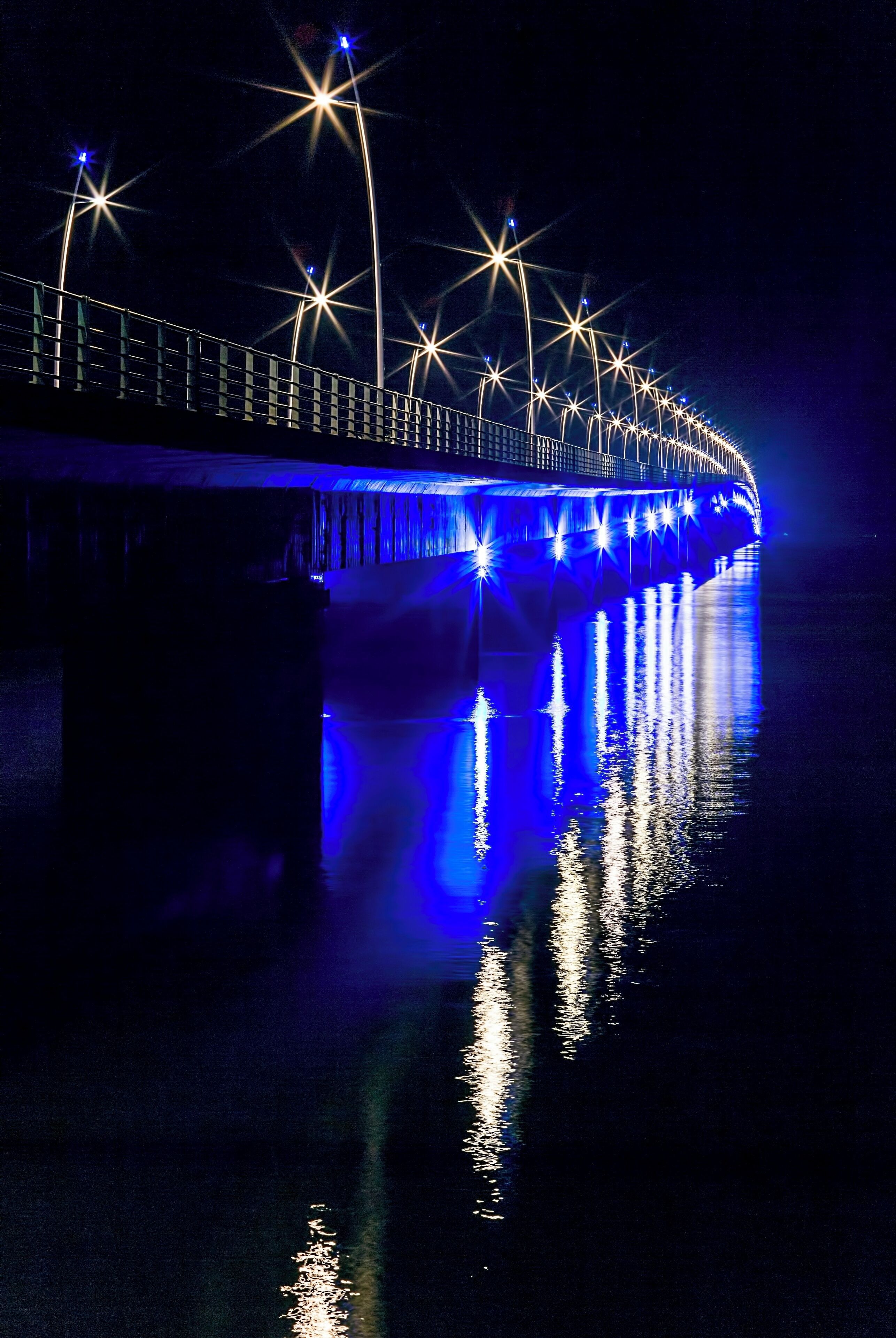 A bridge at night with blue and white lights reflecting on the water.