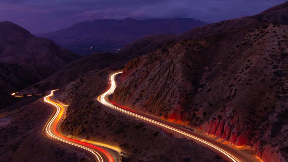 A winding road through a mountainous landscape at twilight with light trails from vehicles.