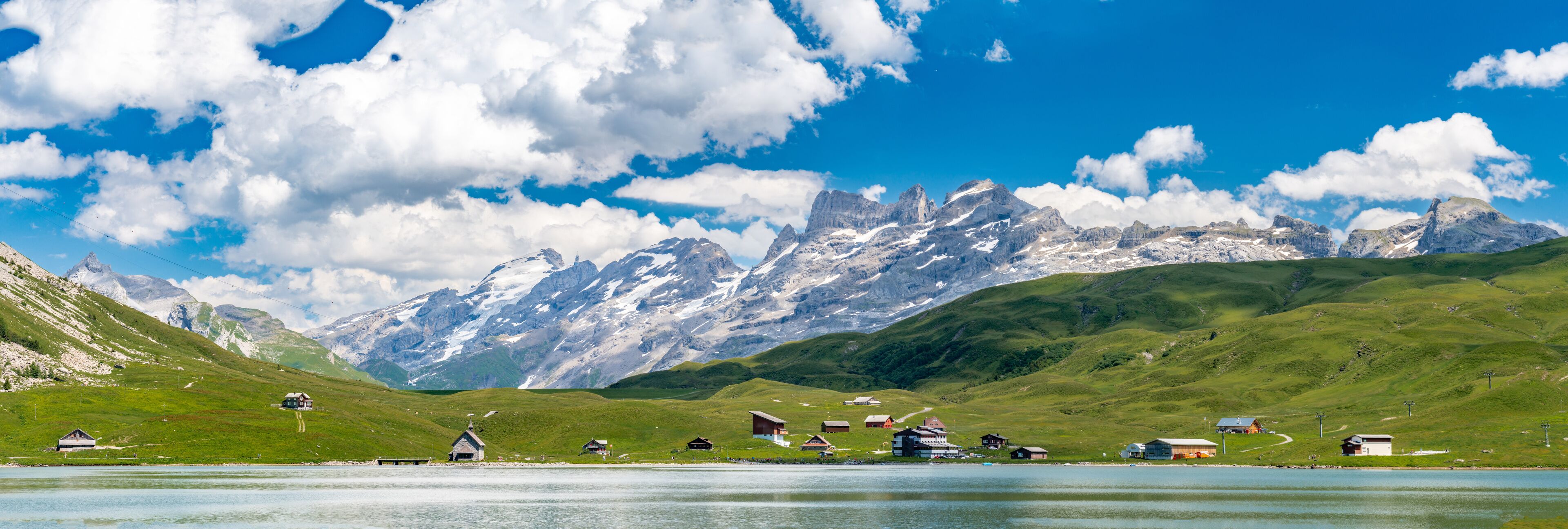 A lake surrounded by green hills and snow-capped mountains under a clear blue sky.