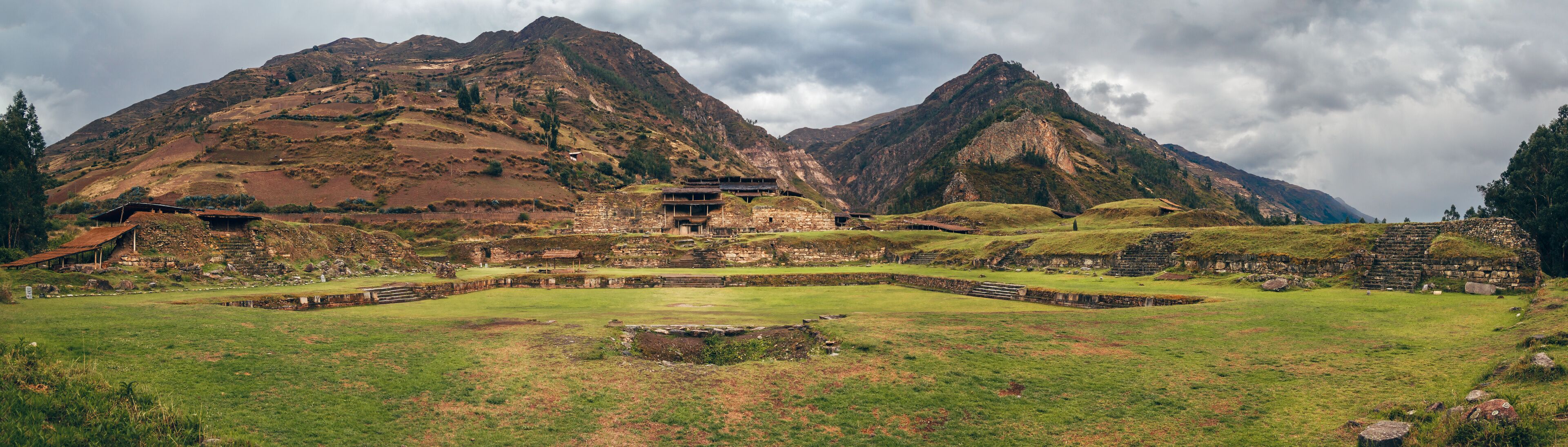 A historical site with ancient ruins and a large open field in front of a mountain range.