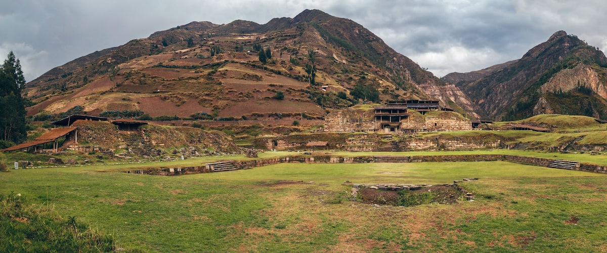 A historical site with ancient ruins and a large open field in front of a mountain range.