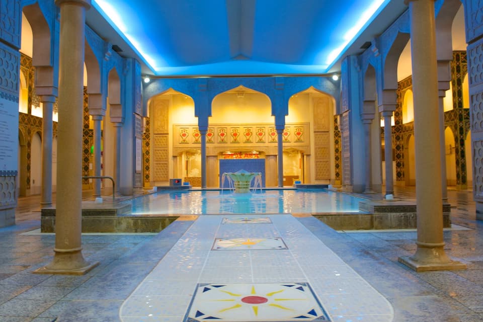 Een binnenzwembad met versierde bogen en zuilen, een betegelde vloer met geometrische patronen en een blauw verlicht plafond.