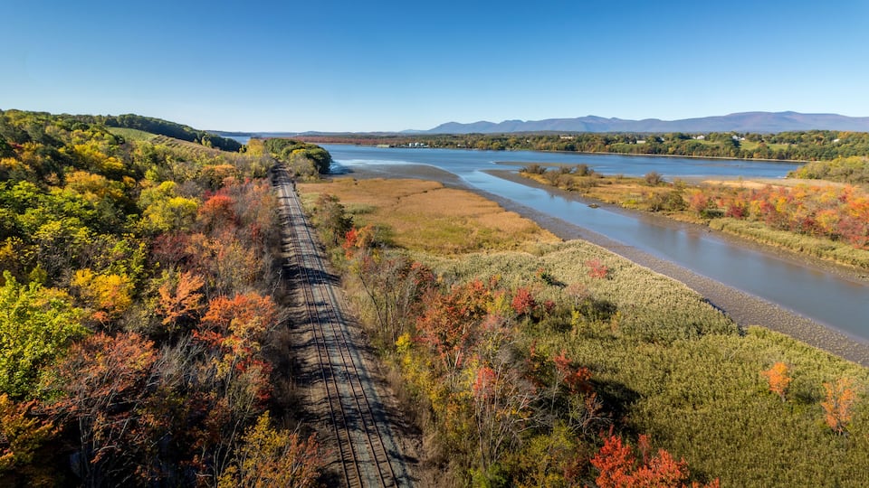 A train track through a forested area with a river and mountains in the background.