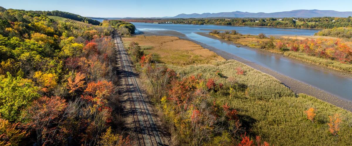 A train track through a forested area with a river and mountains in the background.