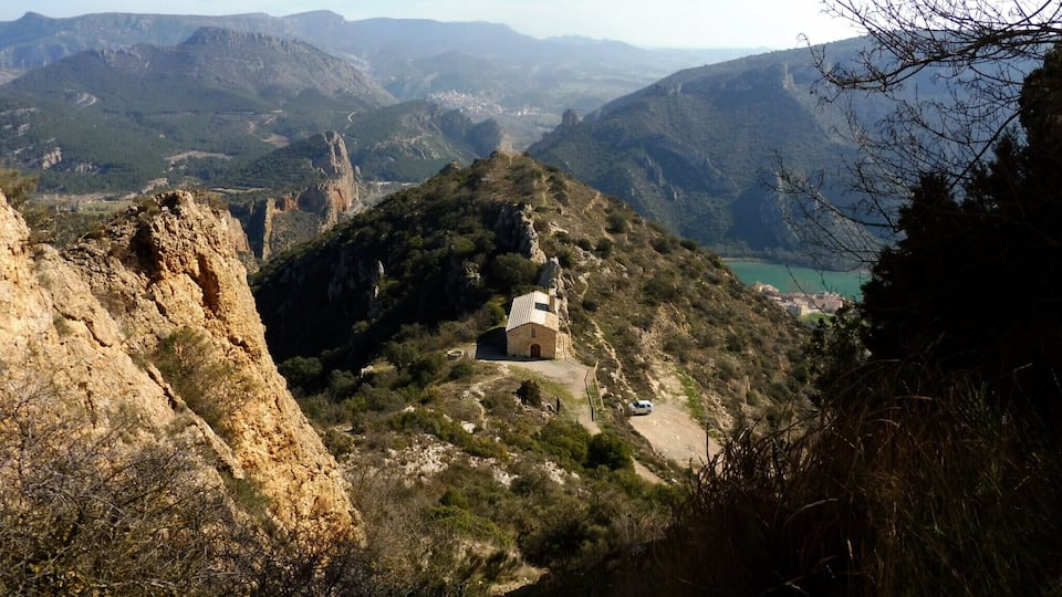 A mountainous landscape with a small building on a hill, surrounded by rocky terrain and sparse vegetation.