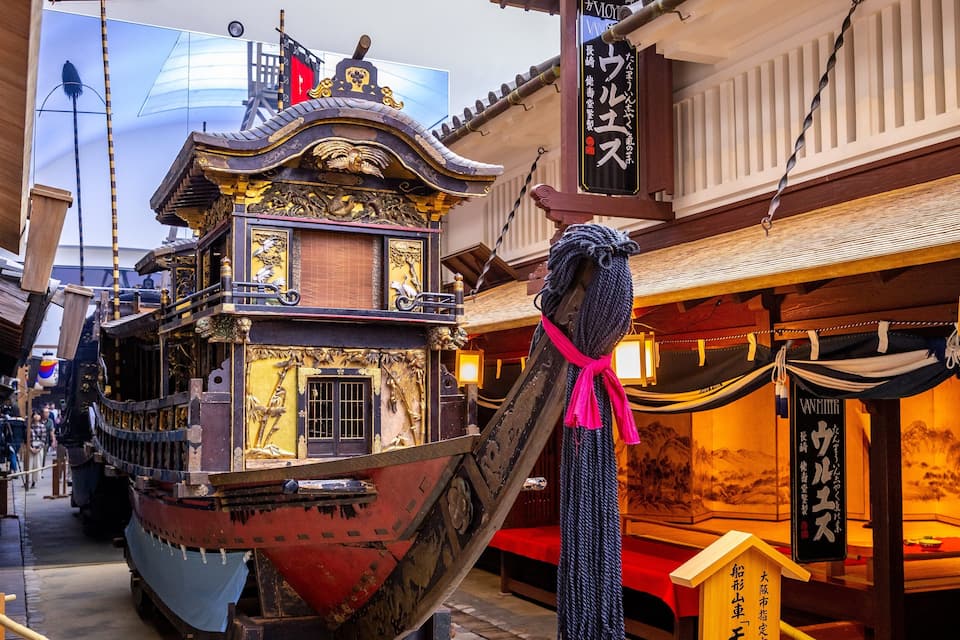Een traditionele houten boot met ingewikkelde houtsnijwerken en een rode romp, afgemeerd in een smalle steeg met traditionele Aziatische architectuur.