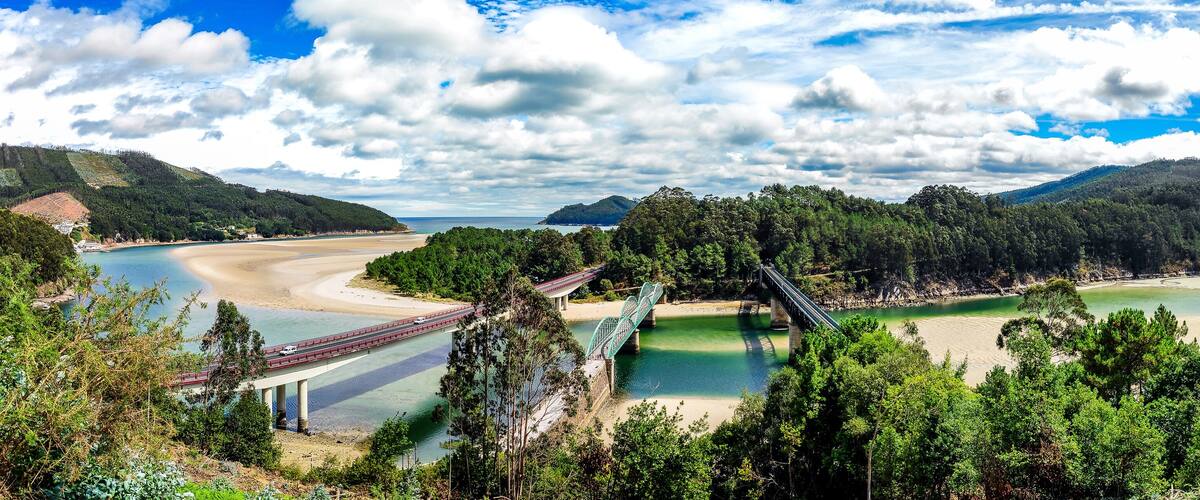 A coastal landscape with a bridge, beach, and forested hills.