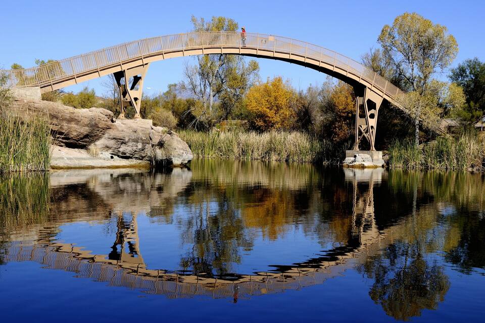 A wooden bridge over a calm river with clear reflections, surrounded by trees and rocks.