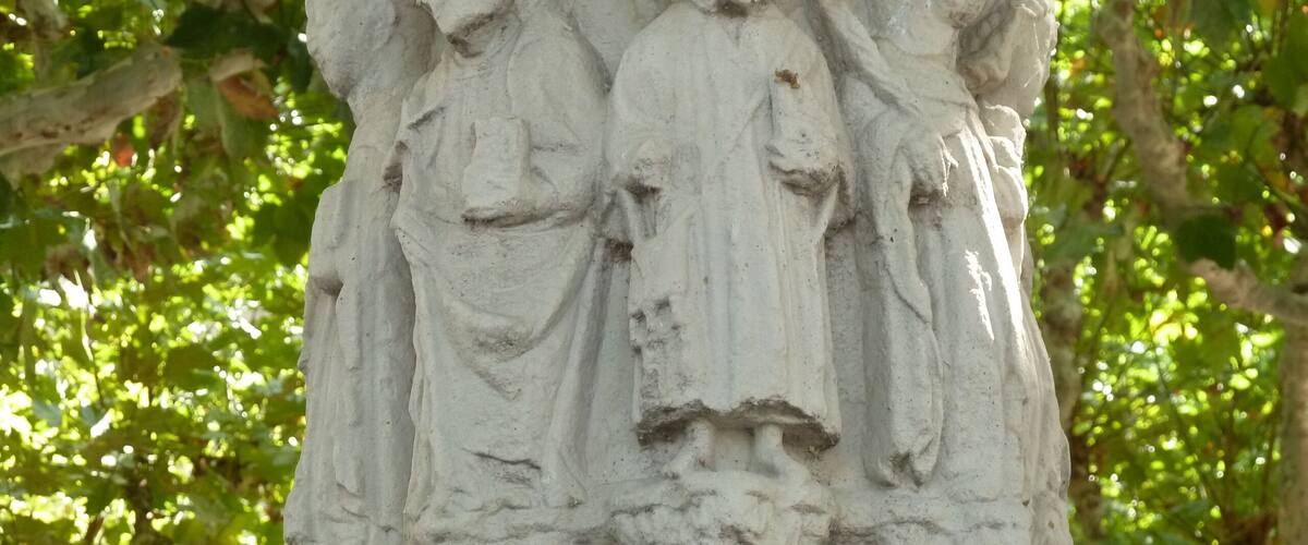 A stone sculpture of three figures with flowing robes, set against a backdrop of green foliage.