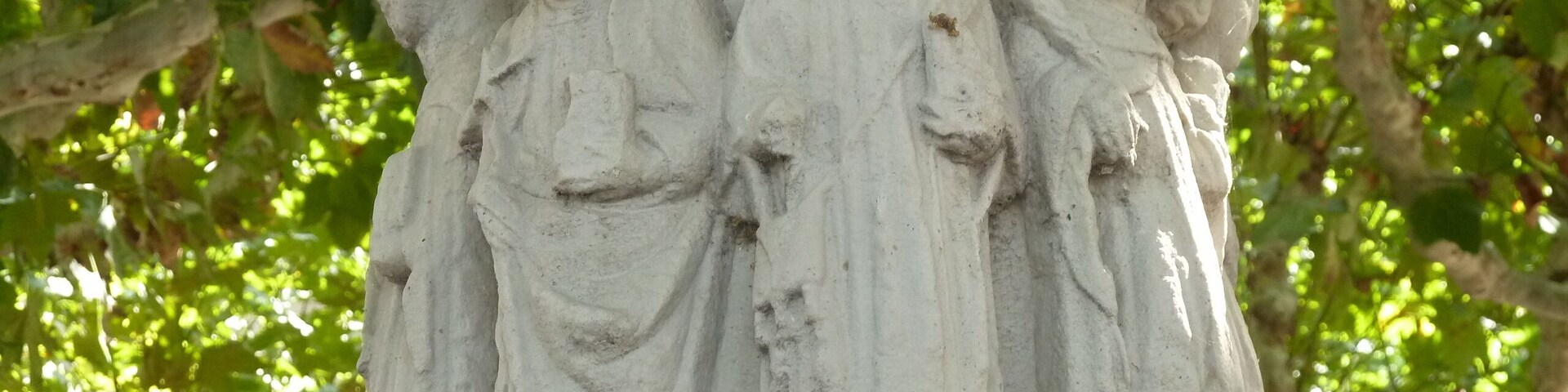 Una escultura de piedra con tres figuras vestidas con túnicas, frente a un fondo de follaje verde.