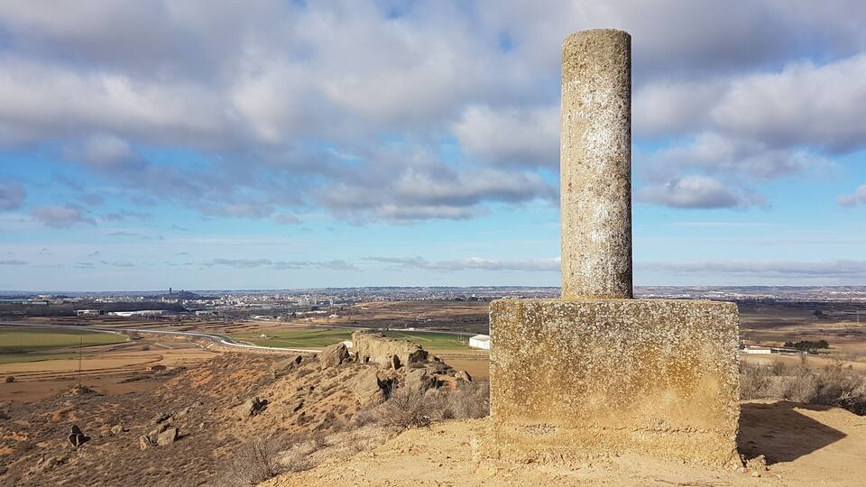 A stone monument with a cylindrical column, surrounded by a hilly landscape and a distant cityscape under a partly cloudy sky.