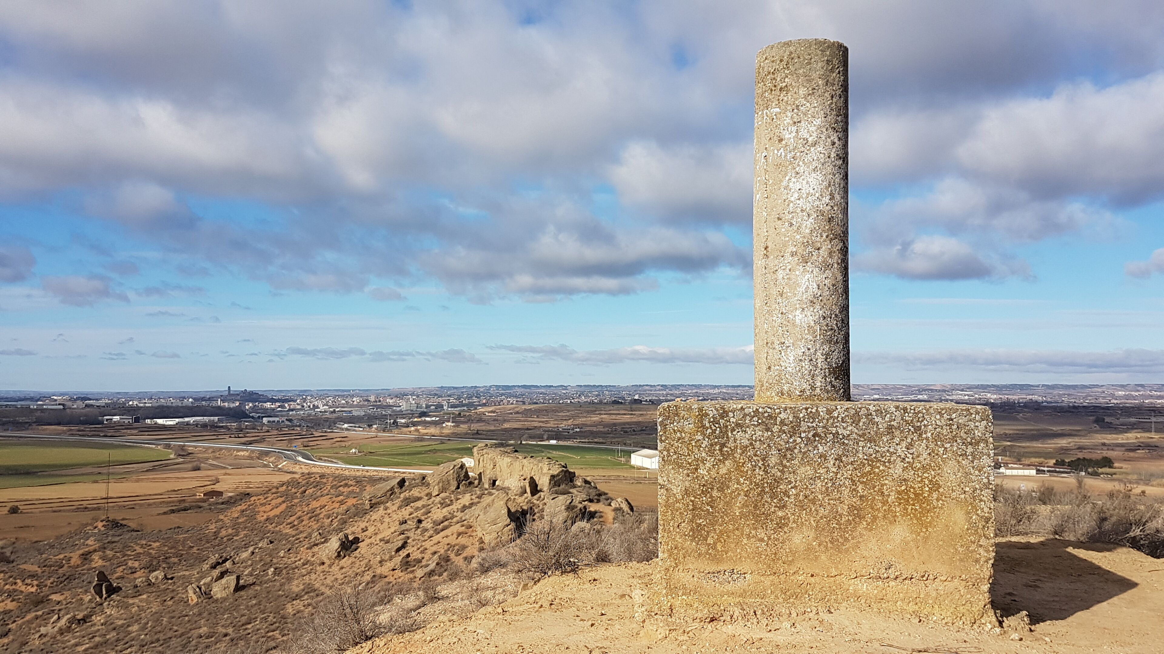 A stone monument with a cylindrical column, surrounded by a hilly landscape and a distant cityscape under a partly cloudy sky.