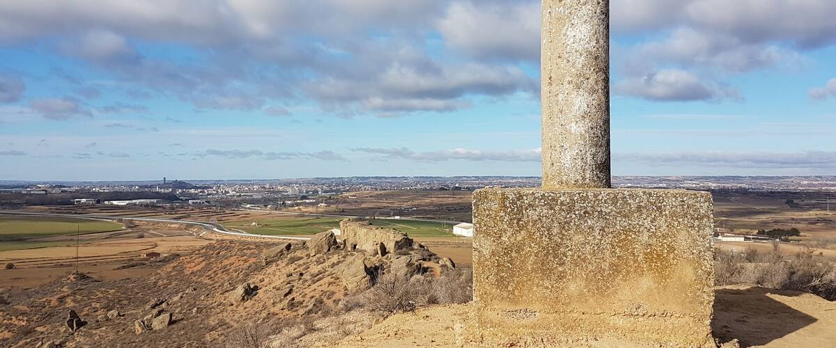 A stone monument with a cylindrical column, surrounded by a hilly landscape and a distant cityscape under a partly cloudy sky.