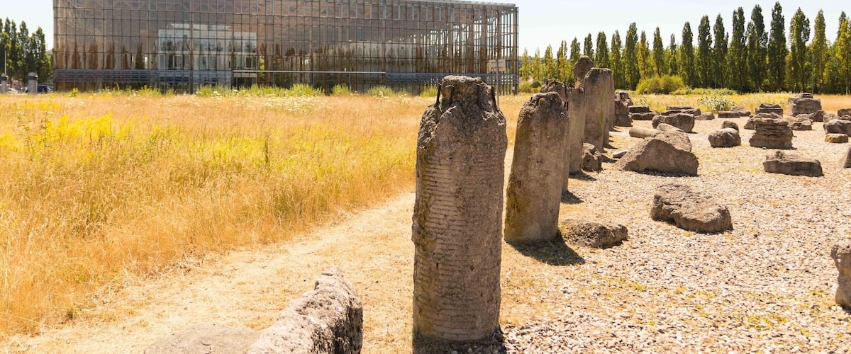 A modern building with a glass facade, surrounded by a field of tall grass and ancient stone pillars.