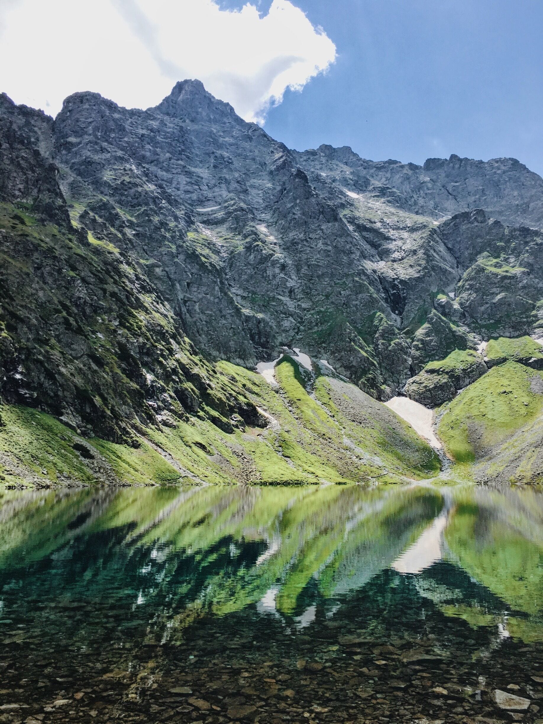 A mountain lake with clear reflections, surrounded by rugged mountains and green vegetation.