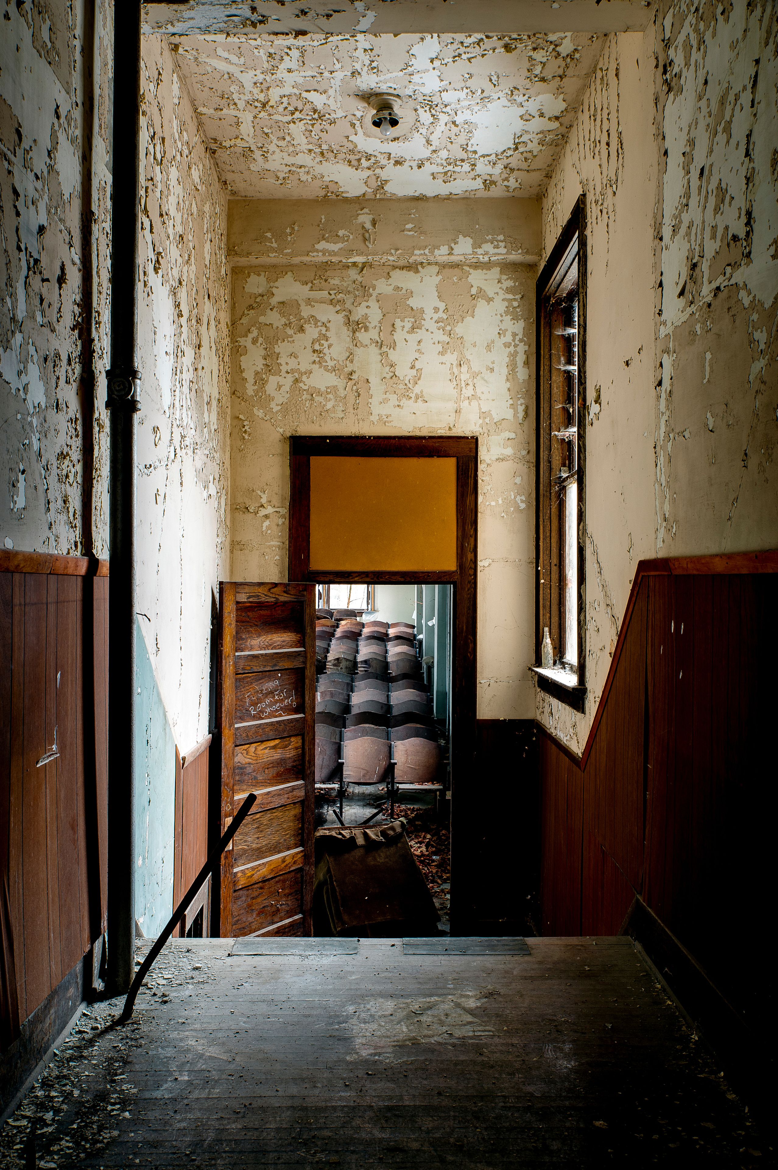 Una habitació deteriorada amb pintura descascarada, una escala de fusta que condueix a una porta i una finestra a la dreta.