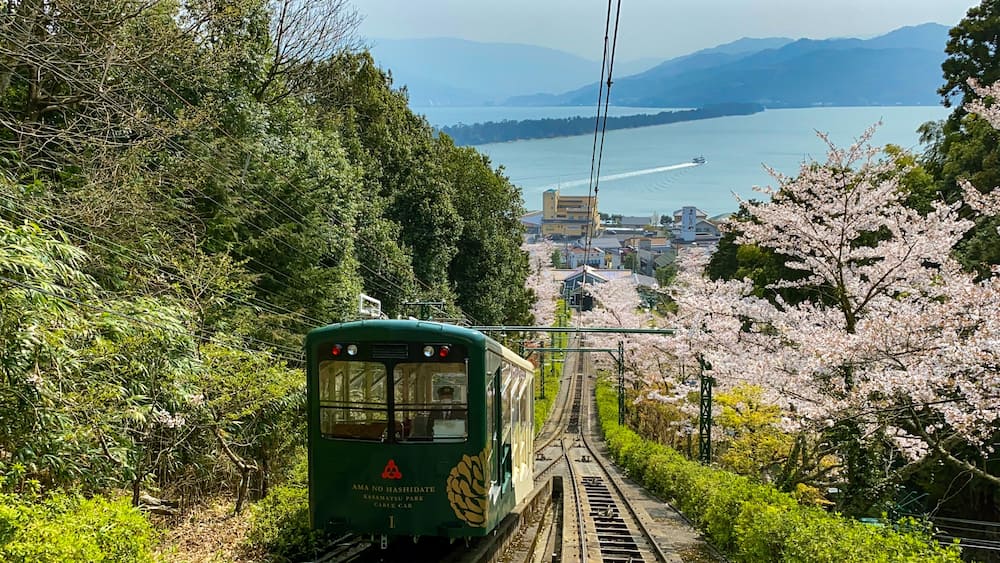 Eine grüne Kabseilbahn fährt auf einer Strecke entlang, umgeben von Kirschblüten, mit Blick auf einen See und Berge in der Ferne.