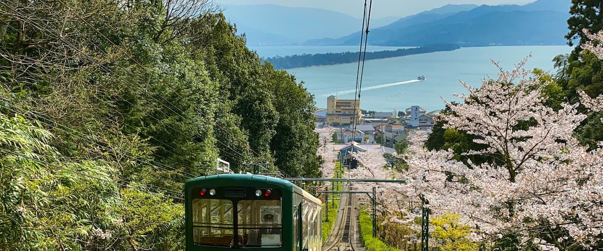 A green cable car ascending a track surrounded by cherry blossoms and a distant view of a lake and mountains.