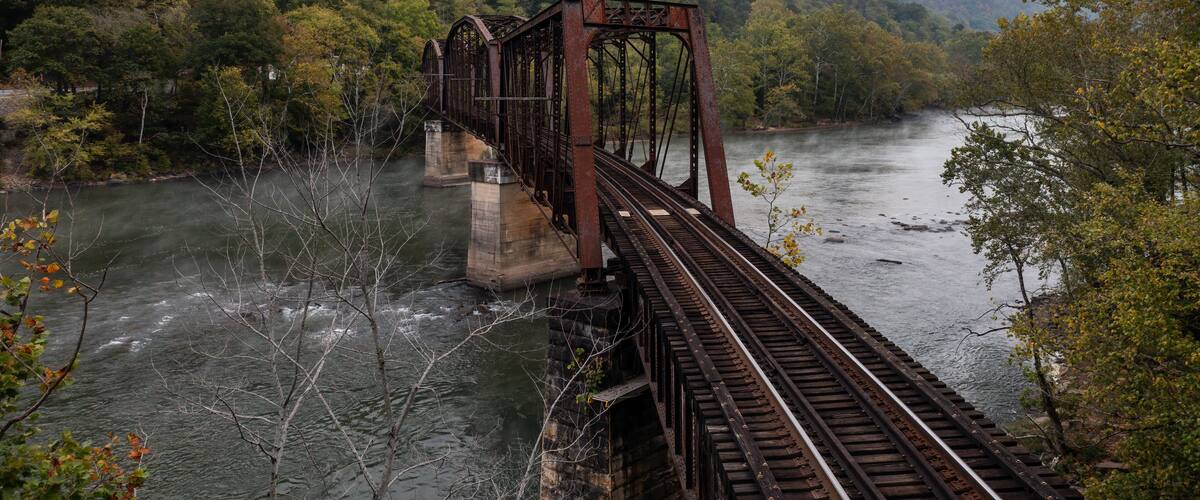 A rusted railway bridge over a river surrounded by dense forest and mist-covered mountains.