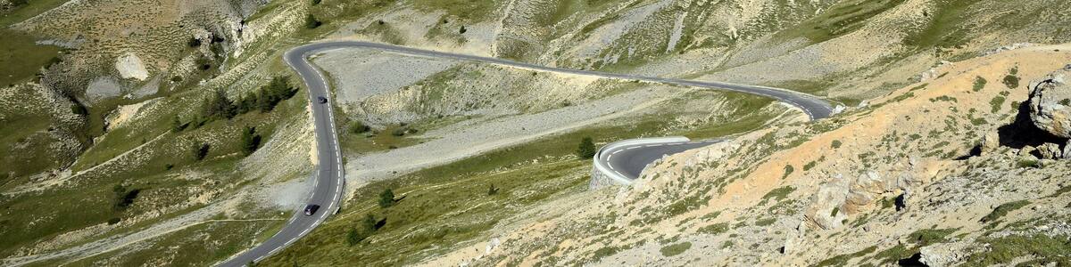 Une route de montagne sinueuse traverse un paysage verdoyant et accidenté, avec des pics imposants et un ciel dégagé.