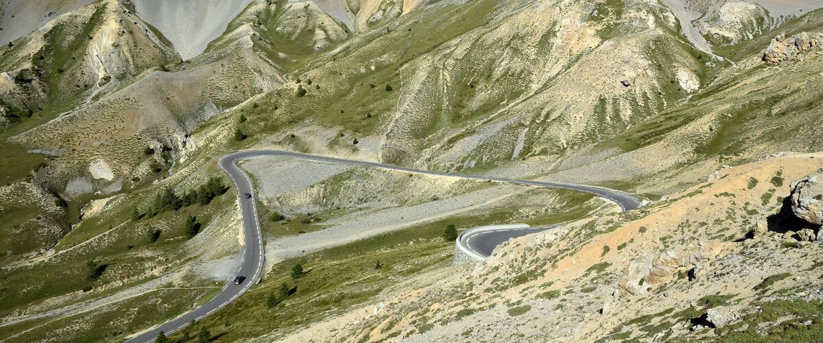 A winding mountain road through a rugged, green landscape with prominent peaks and a clear sky.