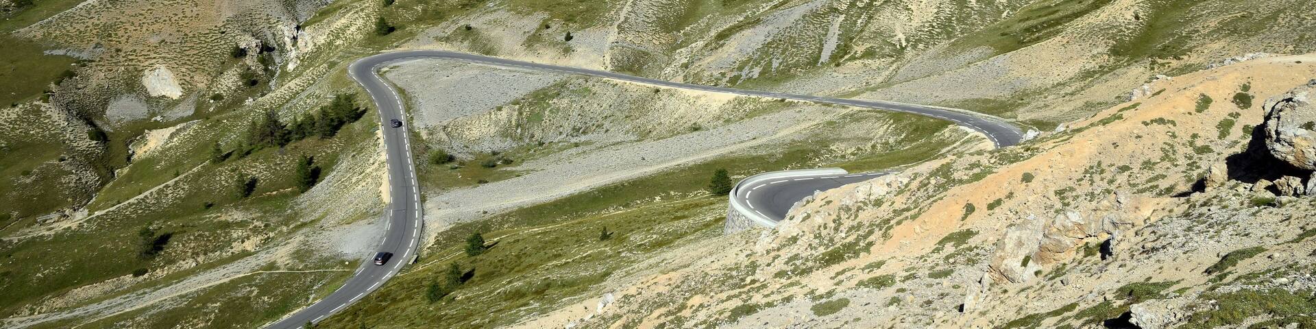 A winding mountain road through a rugged, green landscape with prominent peaks and a clear sky.