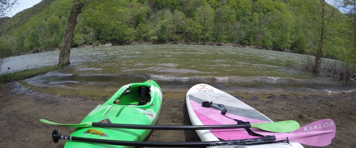 Two kayaks, one green and one white, on a muddy shore with a river and a forested hill in the background.