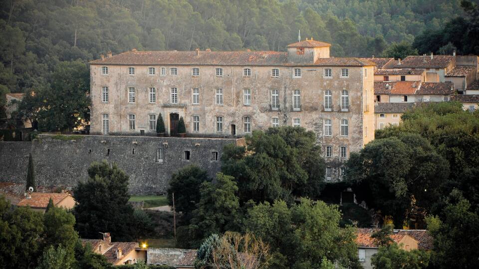 A historic stone building with a tiled roof, surrounded by lush greenery and a stone wall.