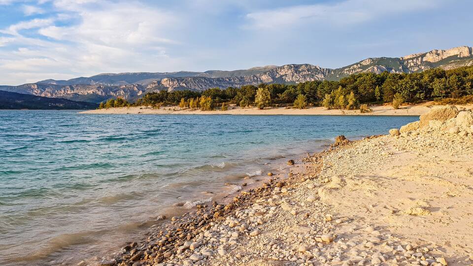 A pebble beach with clear water, a distant mountain range, and sparse trees.