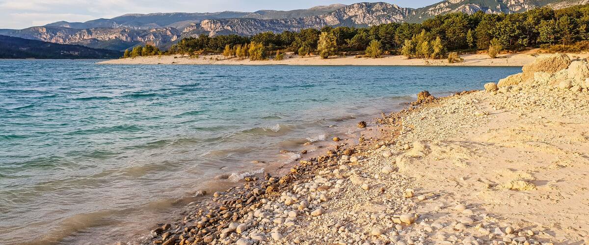 A pebble beach with clear water, a distant mountain range, and sparse trees.