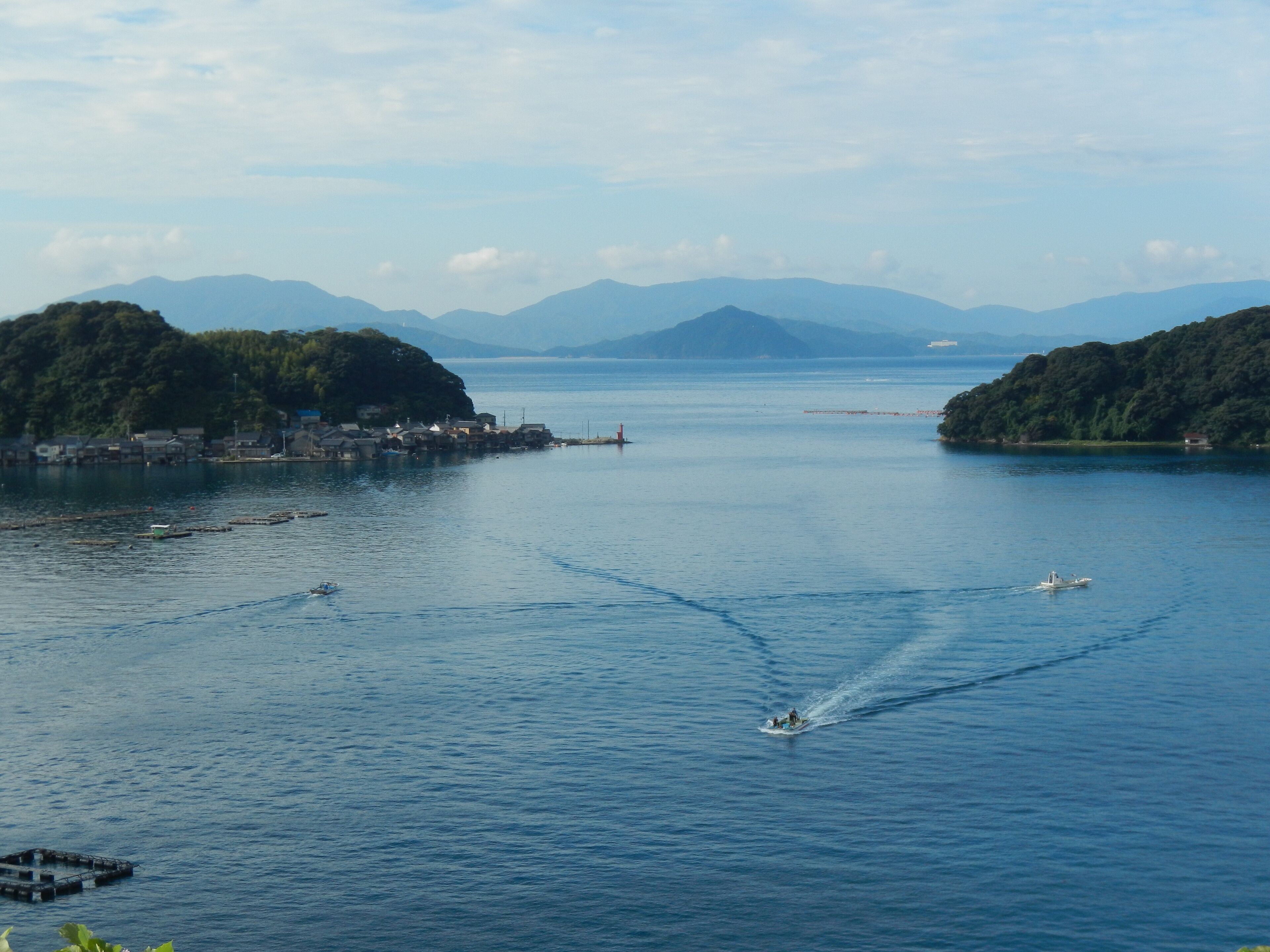 A calm sea with a boat creating a wake, surrounded by hills and a small settlement.