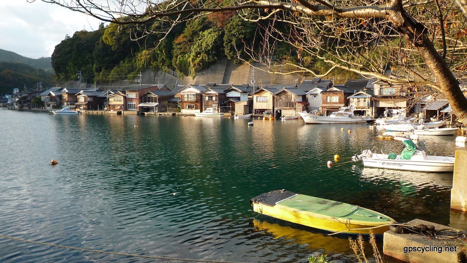 A calm river with boats docked along the shore, surrounded by traditional wooden houses and a forested hillside.