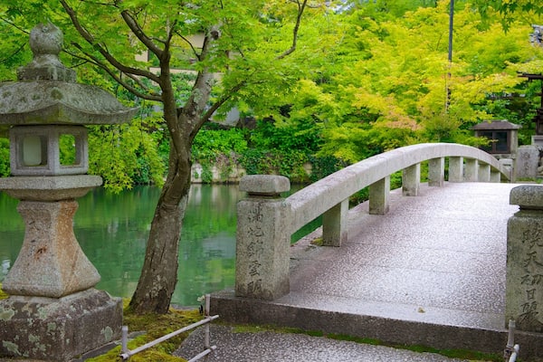 Uma ponte de pedra sobre um rio calmo, cercada por vegetação exuberante e arquitetura japonesa tradicional.