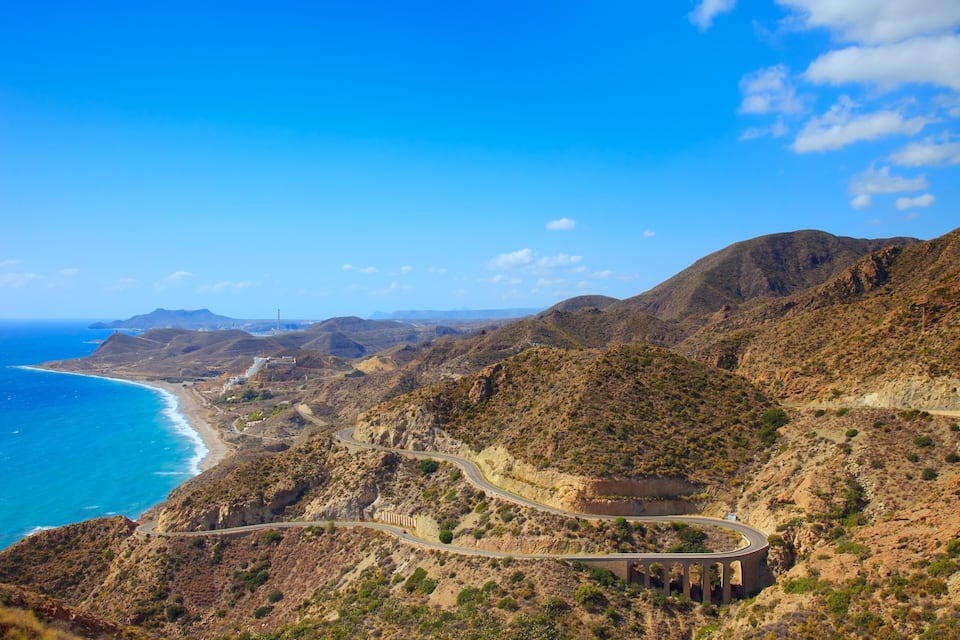 A coastal road winding through a mountainous landscape with a beach and ocean in the background.