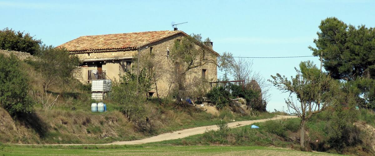 A rustic stone house with a tiled roof, surrounded by a green field and trees.
