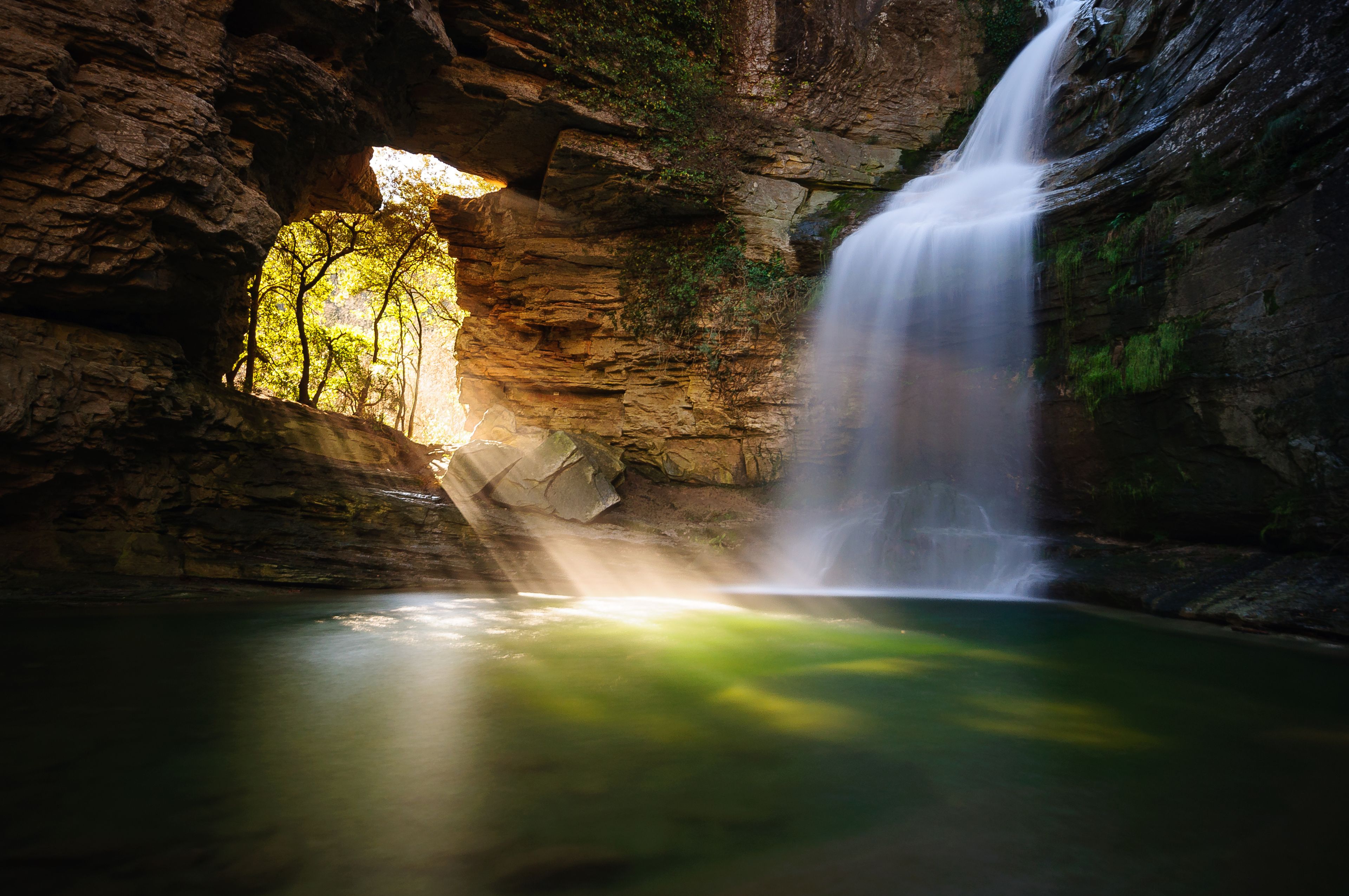 A waterfall cascading into a pool, surrounded by rocky cliffs with a cave entrance.
