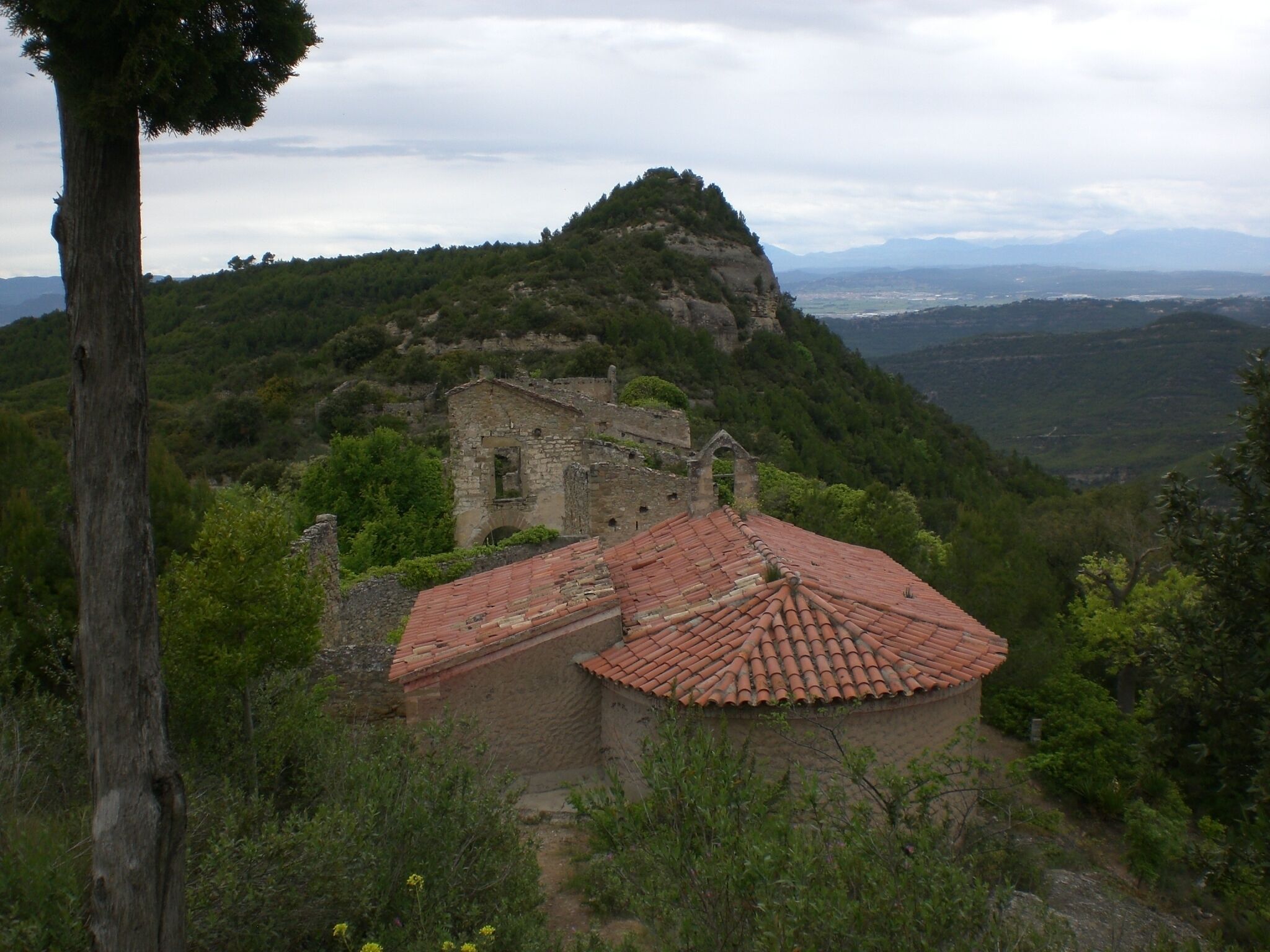 A stone church with a red-tiled roof surrounded by lush greenery and mountains in the background.