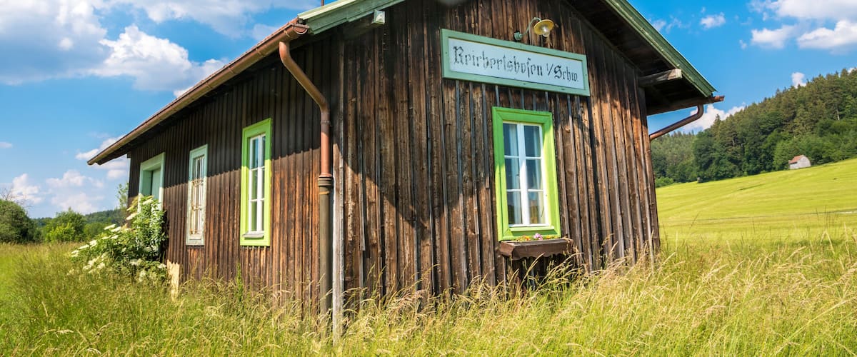 A wooden cabin with green trim and a sign that reads "Rietberleibnig 75b10" in a grassy field with a clear blue sky.