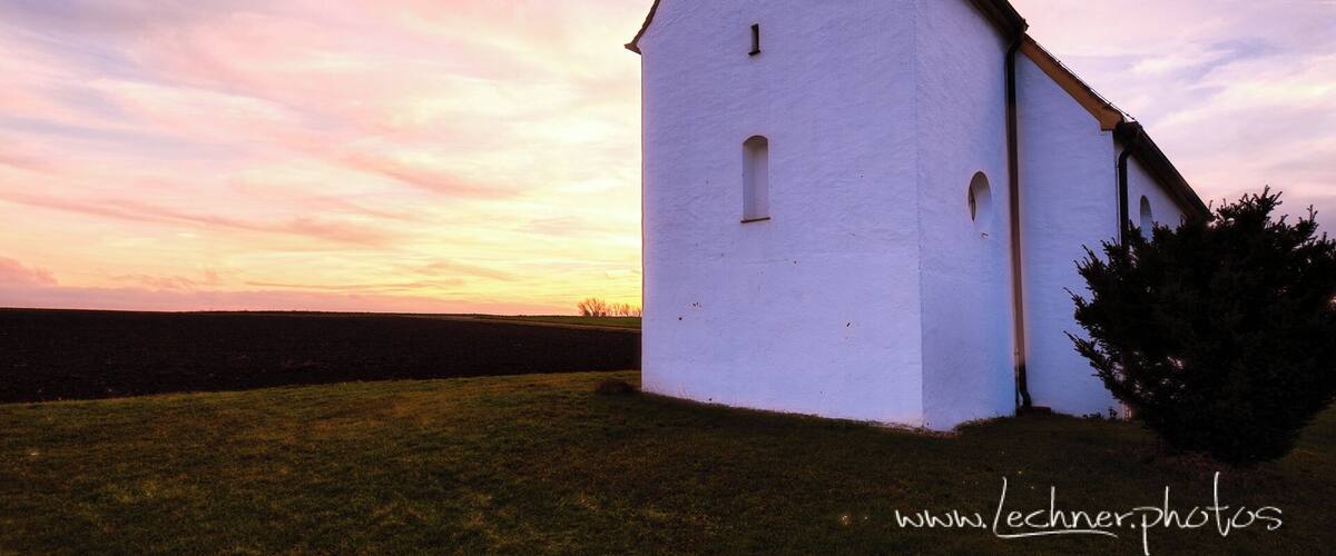 A white church with a black steeple and a clock tower, set against a sunset sky.